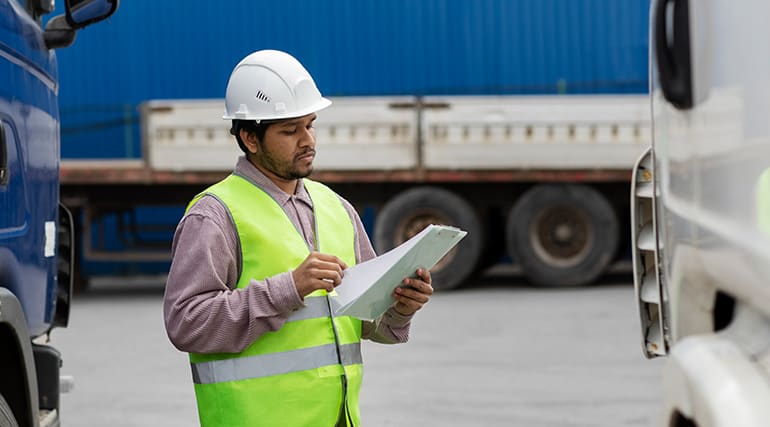 Freight worker in hi-vis vest and hard hat checking delivery paperwork between parked trucks