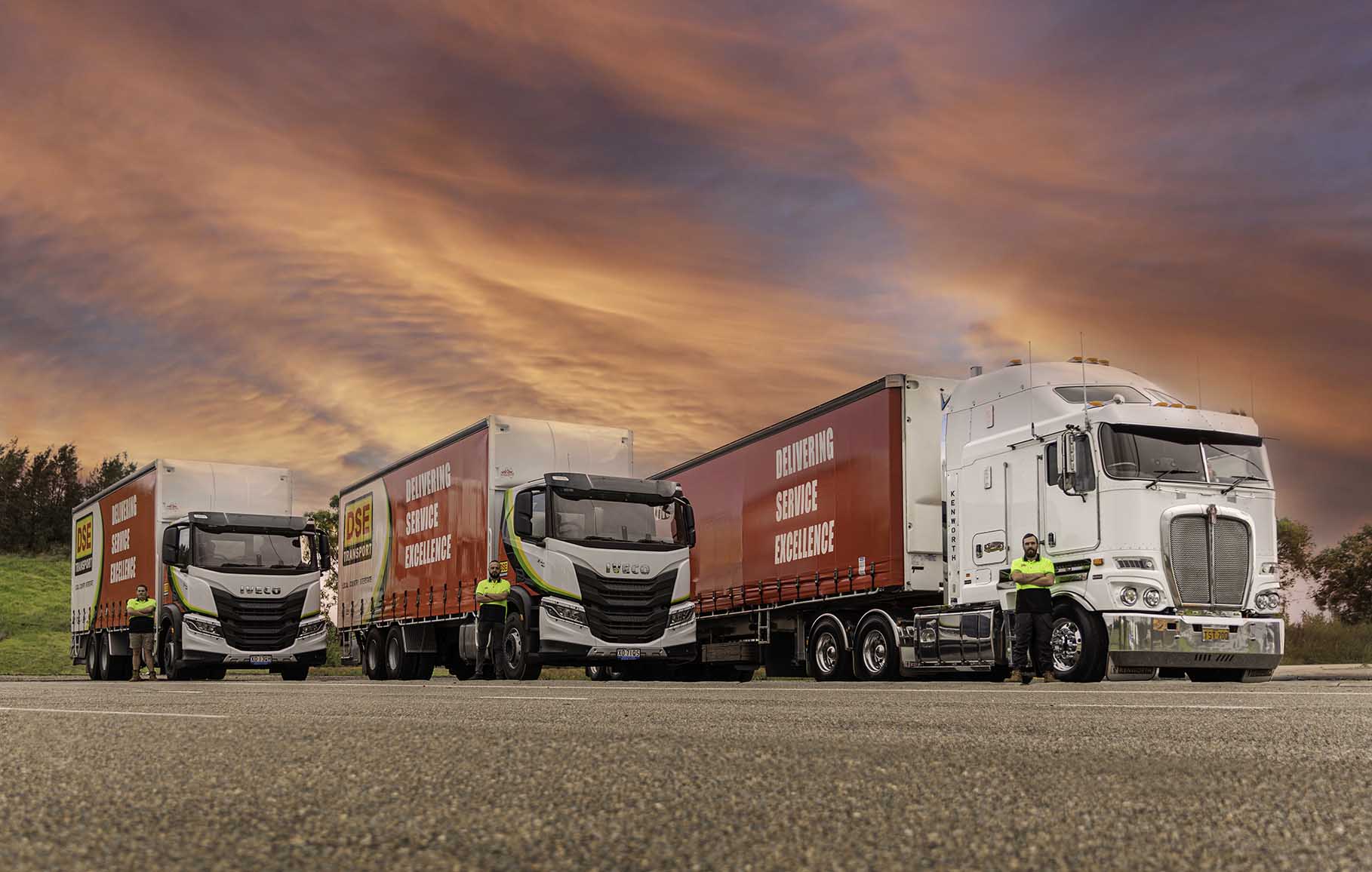 DSE Transport drivers standing beside their rigid trucks and Kenworth semi at dusk
